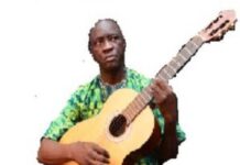 La magie de Noël prend vie au Centre de Landja avec le CREA Monsieur Gervais Lakosso avec une guitard à la main en train de chanter lakosso-guitard Voyagez à travers les Contes et Chants envoûtants de Centrafrique avec Gervais LAKOSSO